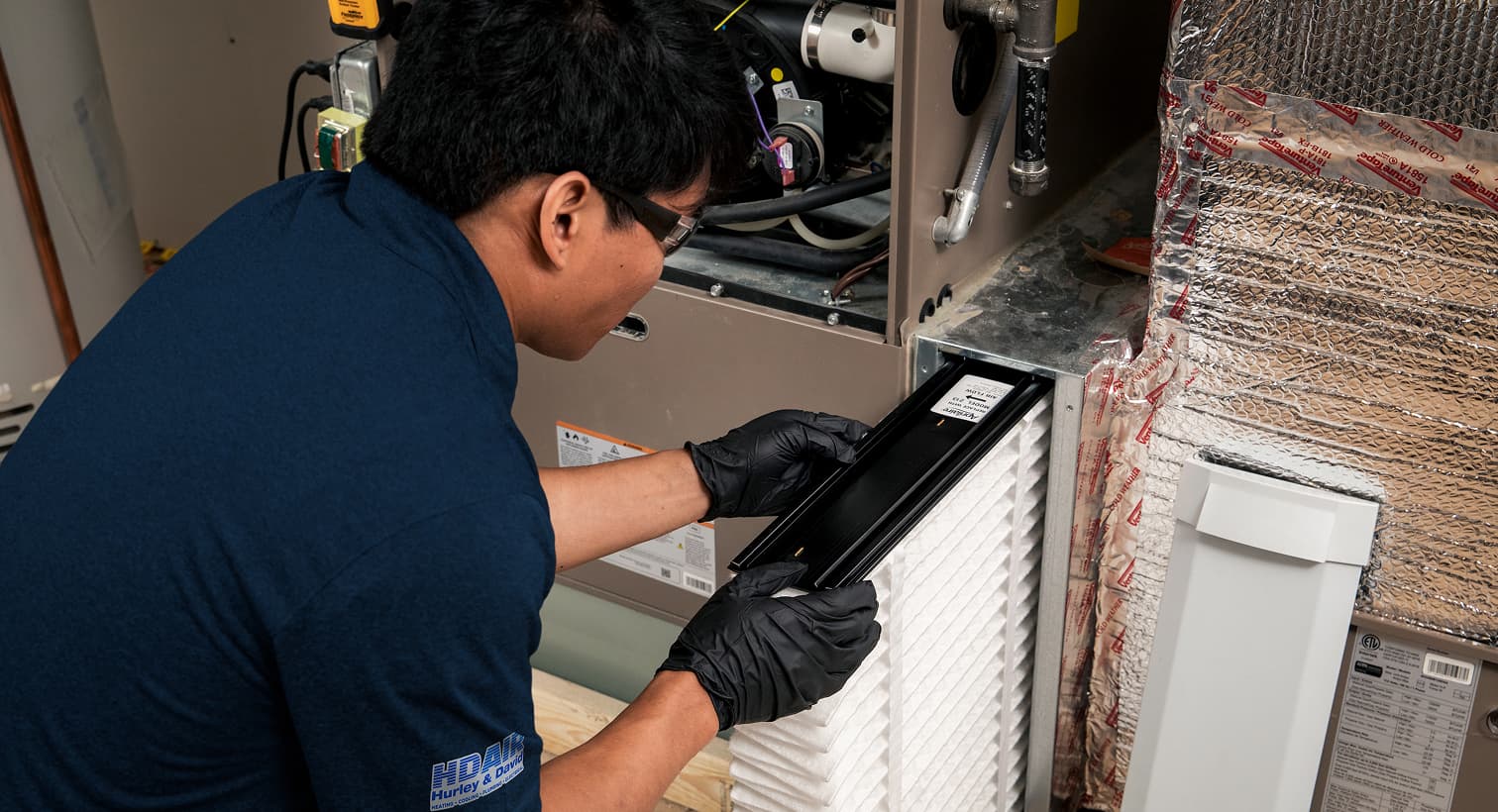 A professional Hurley and David HVAC technician replacing a clean pleated air filter in an indoor air handler unit during a routine AC maintenance tune-up.