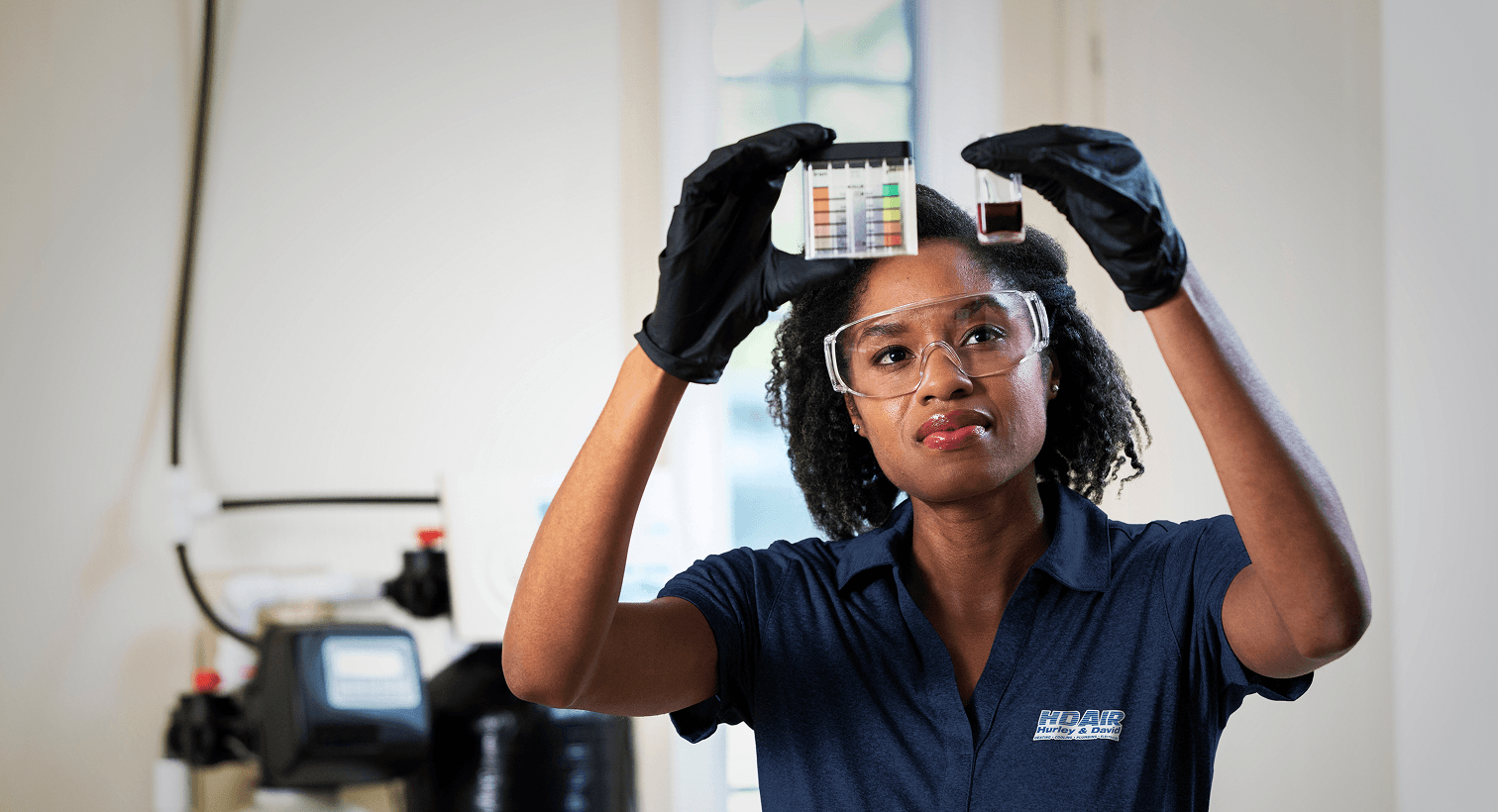 A HD Air technician in a blue uniform and safety gear compares a water sample to a testing chart in front of a water softener system.